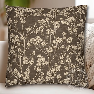 May include: Square decorative pillow with a dark brown background and a cream-colored floral pattern. The pillow has a textured appearance and is likely made of fabric. The design features stylized branches with small flower clusters, creating a natural, botanical theme.