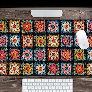 May include: A rectangular mousepad with a black background and a colorful, crocheted floral pattern. The pattern features squares with flowers in shades of orange, pink, yellow, and blue. A white computer mouse rests on the pad, with a keyboard below.