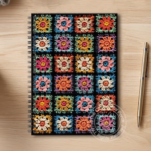 May include: A spiral-bound notebook with a black cover featuring a colorful crochet floral pattern. The design includes squares of various colors, including pink, orange, yellow, and blue. A gold pen rests beside the notebook on a wooden surface.