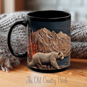 May include: Black ceramic mug featuring a detailed illustration of a brown bear walking in front of a mountain range and a forest. The mug has the text "The Old Country Porch".