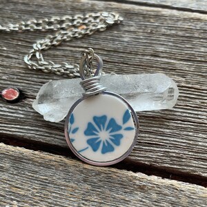 Puede incluir: Un colgante circular que presenta un fragmento de cer&aacute;mica blanca con un patr&oacute;n floral azul, envuelto en un alambre de tono plateado. El colgante cuelga de una cadena de metal plateado, colocado frente a un cristal de cuarzo claro y alargado.