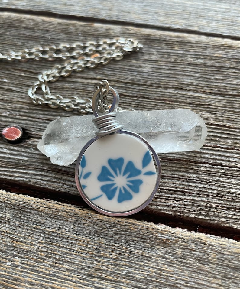 Puede incluir: Un colgante circular que presenta un fragmento de cer&aacute;mica blanca con un patr&oacute;n floral azul, envuelto en un alambre de tono plateado. El colgante cuelga de una cadena de metal plateado, colocado frente a un cristal de cuarzo claro y alargado.