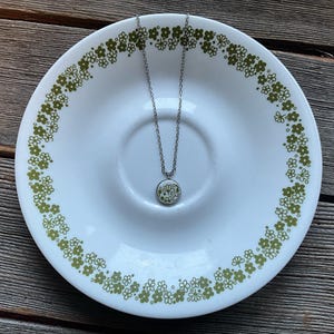 May include: A silver chain necklace with a round pendant featuring a white and green floral design. The necklace is displayed on a white plate with a green floral pattern.