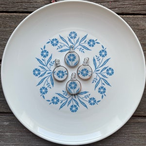 Puede incluir: Cuatro colgantes circulares hechos de cer&aacute;mica blanca con un patr&oacute;n floral azul, cada uno con una anilla de alambre plateado en la parte superior. Los colgantes est&aacute;n dispuestos en grupo, mostrando un dise&ntilde;o bot&aacute;nico consistente en cada pieza.