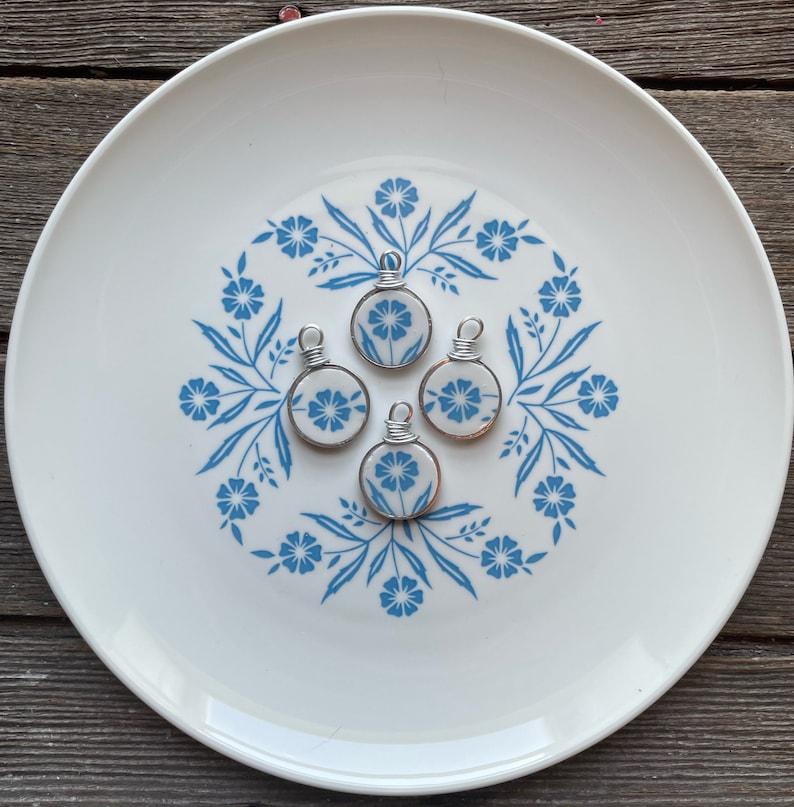 Puede incluir: Cuatro colgantes circulares hechos de cer&aacute;mica blanca con un patr&oacute;n floral azul, cada uno con una anilla de alambre plateado en la parte superior. Los colgantes est&aacute;n dispuestos en grupo, mostrando un dise&ntilde;o bot&aacute;nico consistente en cada pieza.