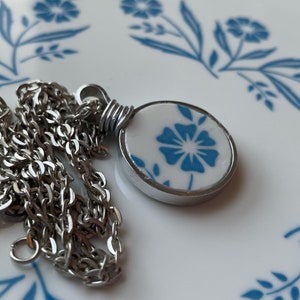 Puede incluir: Un collar de cadena de metal plateado con un colgante circular. El colgante tiene una incrustaci&oacute;n blanca similar a la cer&aacute;mica decorada con un patr&oacute;n floral azul, asegurada por una anilla de alambre plateado en el marco redondo.