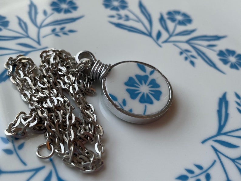 Puede incluir: Un collar de cadena de metal plateado con un colgante circular. El colgante tiene una incrustaci&oacute;n blanca similar a la cer&aacute;mica decorada con un patr&oacute;n floral azul, asegurada por una anilla de alambre plateado en el marco redondo.