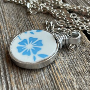 Puede incluir: Un collar con colgante circular que presenta una pieza de cer&aacute;mica blanca con un patr&oacute;n floral azul. El colgante est&aacute; engastado en un bisel de metal plateado, asegurado con alambre plateado y unido a una cadena de eslabones plateada.
