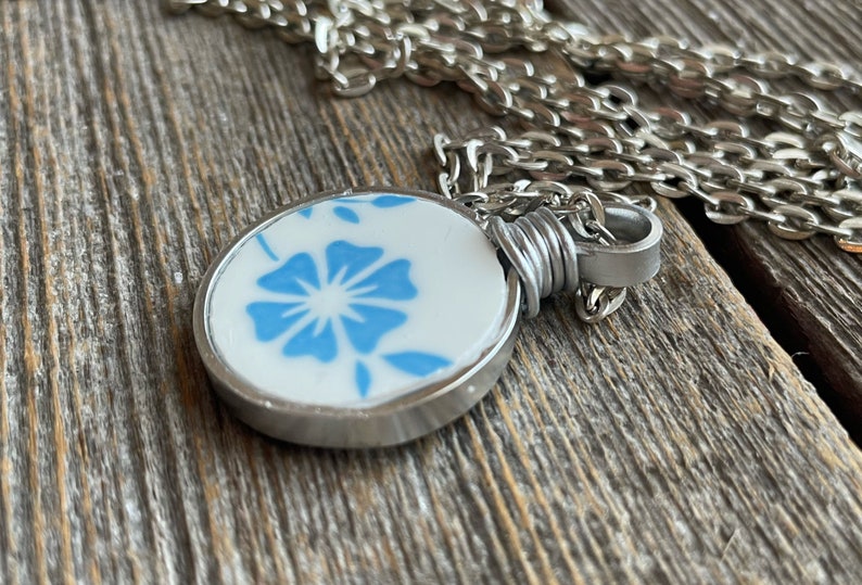 Puede incluir: Un collar con colgante circular que presenta una pieza de cer&aacute;mica blanca con un patr&oacute;n floral azul. El colgante est&aacute; engastado en un bisel de metal plateado, asegurado con alambre plateado y unido a una cadena de eslabones plateada.