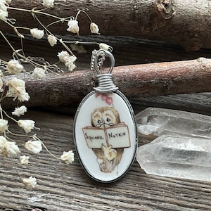 Può includere: Una collana d'argento con un ciondolo ovale che presenta un gufo dei cartoni animati che legge un libro. Il libro dice "Squirrel Nutkin" sulla copertina. Il ciondolo è fatto da un cucchiaio riutilizzato.