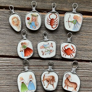 Puede incluir: Una colecci&oacute;n de diez colgantes hechos a mano con signos del zodiaco. Cada colgante tiene una baldosa de cer&aacute;mica blanca con una ilustraci&oacute;n pintada a mano de un s&iacute;mbolo del zodiaco, enmarcada en alambre de plata. Los colgantes est&aacute;n dispuestos sobre una superficie de madera.
