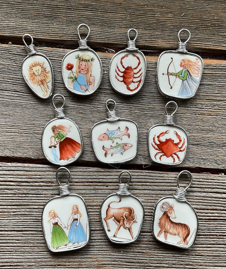 Puede incluir: Una colecci&oacute;n de diez colgantes hechos a mano con signos del zodiaco. Cada colgante tiene una baldosa de cer&aacute;mica blanca con una ilustraci&oacute;n pintada a mano de un s&iacute;mbolo del zodiaco, enmarcada en alambre de plata. Los colgantes est&aacute;n dispuestos sobre una superficie de madera.