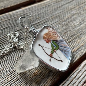Puede incluir: Collar con colgante de plata con un colgante en forma de escudo. El colgante tiene una ilustraci&oacute;n pintada de una figura con arco y flechas. La figura tiene cabello rubio, una camisa verde y una capa azul. Incluye un cristal transparente y una cadena de plata.