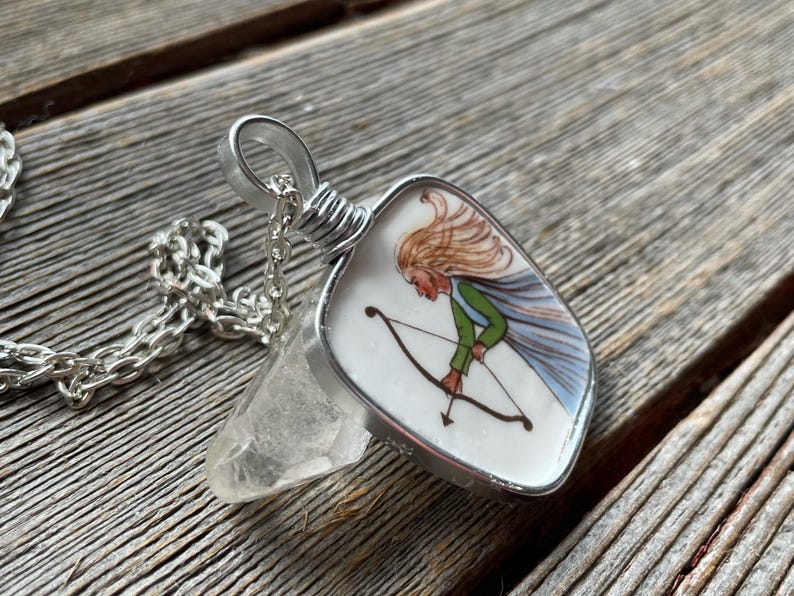 Puede incluir: Collar con colgante de plata con un colgante en forma de escudo. El colgante tiene una ilustraci&oacute;n pintada de una figura con arco y flechas. La figura tiene cabello rubio, una camisa verde y una capa azul. Incluye un cristal transparente y una cadena de plata.