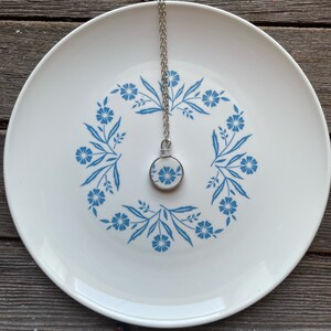 Puede incluir: Un collar de cadena plateada con un colgante circular hecho de un fragmento de cer&aacute;mica blanca con un patr&oacute;n floral azul. El colgante est&aacute; asegurado con alambre plateado y descansa sobre un plato blanco con un dise&ntilde;o floral azul a juego.