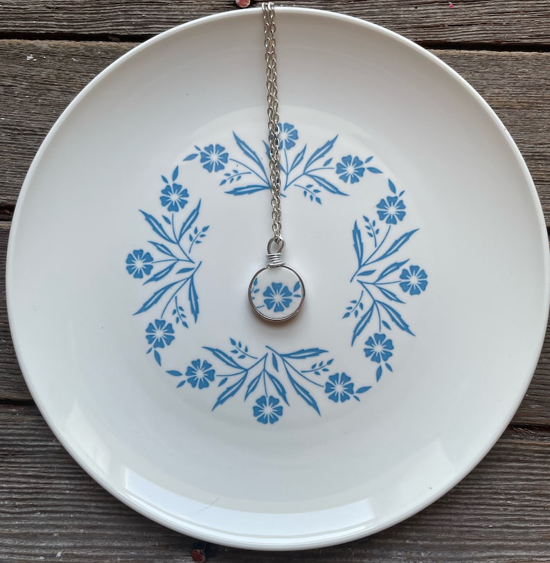 Puede incluir: Un collar de cadena plateada con un colgante circular hecho de un fragmento de cer&aacute;mica blanca con un patr&oacute;n floral azul. El colgante est&aacute; asegurado con alambre plateado y descansa sobre un plato blanco con un dise&ntilde;o floral azul a juego.