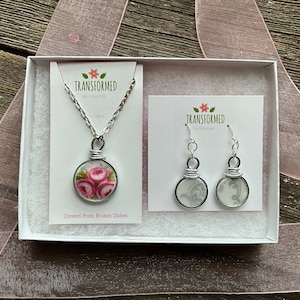 Puede incluir: Una caja de regalo blanca con un collar y pendientes de plata. El collar tiene un colgante hecho de un plato de porcelana vintage con rosas rosas. Los pendientes están hechos de platos de porcelana vintage con un diseño floral gris y blanco. La caja tiene una etiqueta que dice "TRANSFORMED" y "Creado a partir de platos rotos".