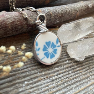 Puede incluir: Un collar con colgante circular que presenta una pieza de cer&aacute;mica blanca con un patr&oacute;n floral azul, engastada en un marco de metal plateado. El colgante est&aacute; unido a una cadena plateada mediante una anilla de alambre enrollado.