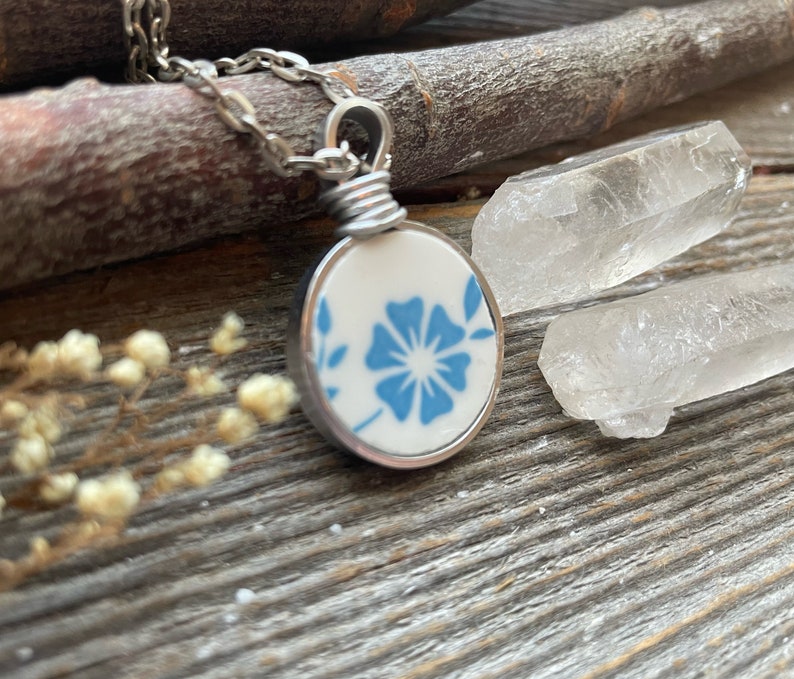 Puede incluir: Un collar con colgante circular que presenta una pieza de cer&aacute;mica blanca con un patr&oacute;n floral azul, engastada en un marco de metal plateado. El colgante est&aacute; unido a una cadena plateada mediante una anilla de alambre enrollado.