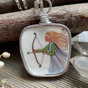 Puede incluir: Un collar con colgante de plata con una baldosa de cer&aacute;mica blanca cuadrada con una ilustraci&oacute;n pintada a mano de una mujer con arco y flecha. La mujer tiene el pelo largo y rubio, una blusa verde y una capa azul. El colgante est&aacute; envuelto en alambre y cuelga de una cadena de plata.