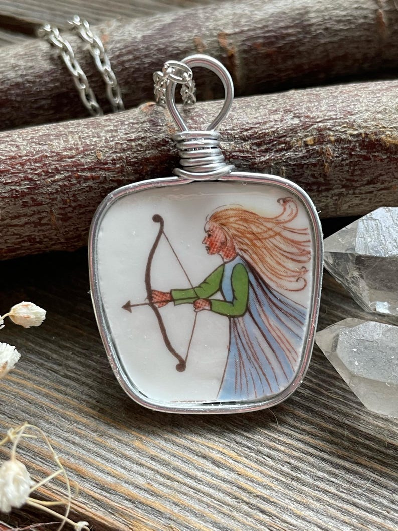 Puede incluir: Un collar con colgante de plata con una baldosa de cer&aacute;mica blanca cuadrada con una ilustraci&oacute;n pintada a mano de una mujer con arco y flecha. La mujer tiene el pelo largo y rubio, una blusa verde y una capa azul. El colgante est&aacute; envuelto en alambre y cuelga de una cadena de plata.
