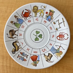 Puede incluir: Un plato de cer&aacute;mica blanco con un dise&ntilde;o del zodiaco. El plato tiene un dise&ntilde;o central de tr&eacute;bol y est&aacute; rodeado de s&iacute;mbolos e ilustraciones del zodiaco en varios colores, incluyendo rojo, dorado y azul. El plato tiene un aspecto ligeramente envejecido.