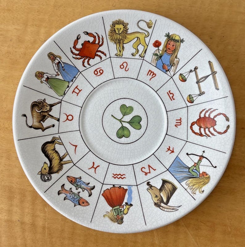 Puede incluir: Un plato de cer&aacute;mica blanco con un dise&ntilde;o del zodiaco. El plato tiene un dise&ntilde;o central de tr&eacute;bol y est&aacute; rodeado de s&iacute;mbolos e ilustraciones del zodiaco en varios colores, incluyendo rojo, dorado y azul. El plato tiene un aspecto ligeramente envejecido.