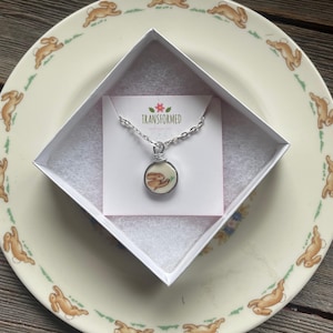 Könnte beinhalten: Eine silberne Kettenkette mit einem runden Anhänger mit einer weißen Porzellan-Hasenillustration. Die Kette befindet sich in einer weißen Schachtel mit rosa Innenausstattung und einer Karte mit der Aufschrift "TRANSFORMED".