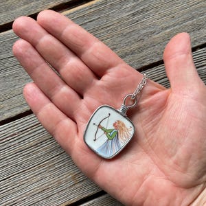 Puede incluir: Un collar con colgante de plata con una imagen cuadrada pintada a mano de una mujer con arco y flecha. La mujer lleva un top verde y un vestido azul. El colgante se sostiene en una mano.