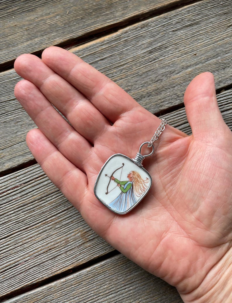 Puede incluir: Un collar con colgante de plata con una imagen cuadrada pintada a mano de una mujer con arco y flecha. La mujer lleva un top verde y un vestido azul. El colgante se sostiene en una mano.