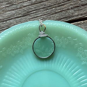 Puede incluir: Un colgante de alambre plateado envuelto con un encanto redondo de vidrio verde claro. El encanto tiene un diseño floral y está sentado en un plato verde claro.