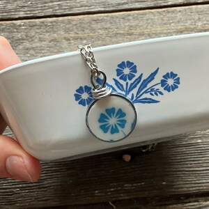Puede incluir: Una fuente de horno rectangular de cer&aacute;mica blanca con un patr&oacute;n floral azul y un colgante circular a juego en una cadena plateada. El colgante muestra un dise&ntilde;o de flor azul dentro de un marco de metal con una anilla de alambre enrollado.