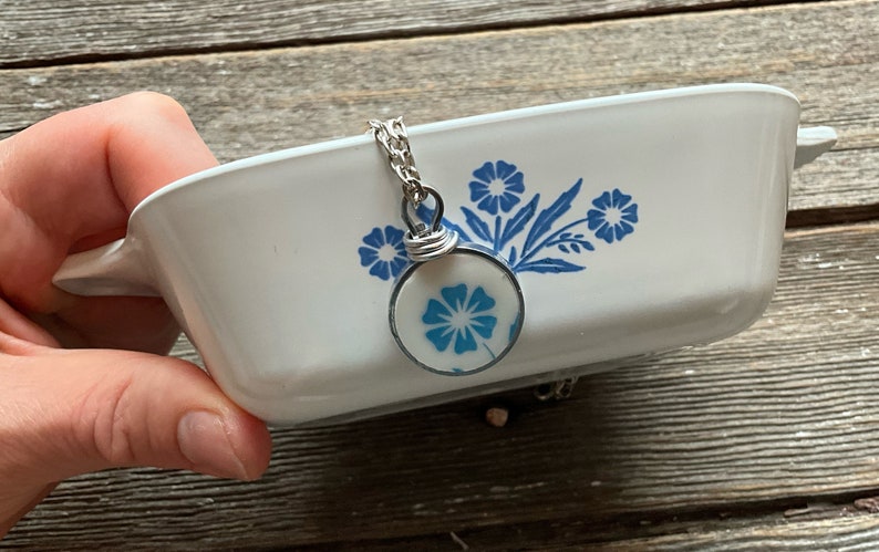 Puede incluir: Una fuente de horno rectangular de cer&aacute;mica blanca con un patr&oacute;n floral azul y un colgante circular a juego en una cadena plateada. El colgante muestra un dise&ntilde;o de flor azul dentro de un marco de metal con una anilla de alambre enrollado.