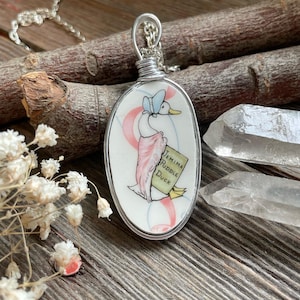 May include: Oval pendant necklace with a hand-painted illustration of Jemima Puddle-Duck. The pendant is set in a silver-toned metal frame and hangs from a delicate chain. The duck is wearing a bonnet and shawl, holding a sign that reads "Jemima Puddle Duck".