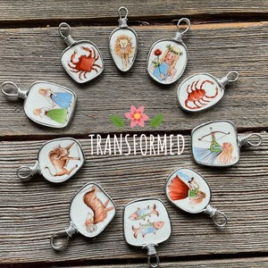 Puede incluir: Una colecci&oacute;n de colgantes de signos del zodiaco hechos a mano. Cada colgante presenta una ilustraci&oacute;n &uacute;nica dentro de un marco de alambre de plata. Los colgantes est&aacute;n dispuestos sobre una superficie de madera, con la palabra "TRANSFORMED" en el centro.