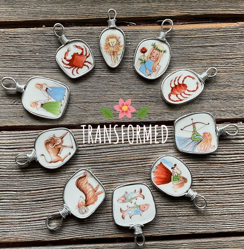 Puede incluir: Una colecci&oacute;n de colgantes de signos del zodiaco hechos a mano. Cada colgante presenta una ilustraci&oacute;n &uacute;nica dentro de un marco de alambre de plata. Los colgantes est&aacute;n dispuestos sobre una superficie de madera, con la palabra "TRANSFORMED" en el centro.