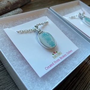 以下が含まれることがあります： 白いギフトボックスに入った2つのネックレス。各ネックレスは、シルバーチェーンにライトブルーのデザインが施されたシルバーペンダントが特徴です。「TRANSFORMED Created From Broken Dishes」の文字がカードに印刷されています。