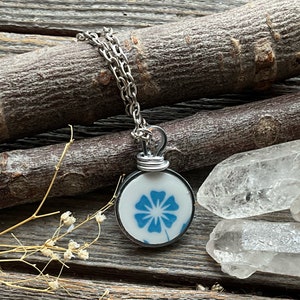 Puede incluir: Un collar con colgante circular que presenta una base de cer&aacute;mica blanca con un patr&oacute;n floral azul. El colgante est&aacute; asegurado con un alambre de tono plateado y cuelga de una cadena de eslabones de metal de tono plateado.
