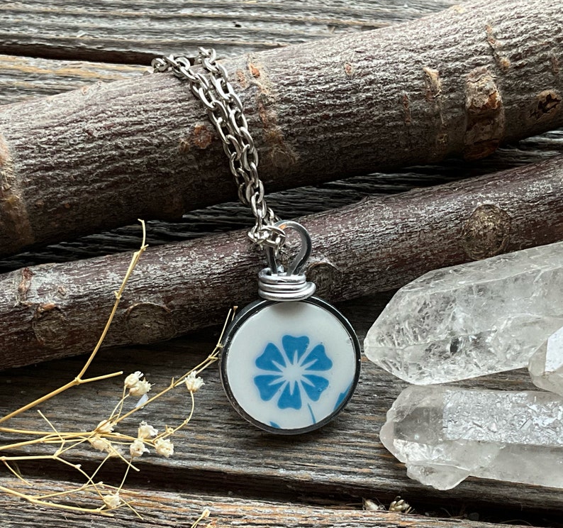 Puede incluir: Un collar con colgante circular que presenta una base de cer&aacute;mica blanca con un patr&oacute;n floral azul. El colgante est&aacute; asegurado con un alambre de tono plateado y cuelga de una cadena de eslabones de metal de tono plateado.