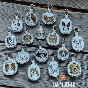May include: A collection of handmade dog pendant necklaces. Each pendant features a different dog breed illustration, encased in a wire frame. The pendants are on a wooden surface. The word "TRANSFORMED" is at the bottom.