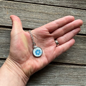 Puede incluir: Un colgante circular con una base de cer&aacute;mica blanca y un patr&oacute;n floral azul, engastado en un marco de metal plateado. El colgante est&aacute; unido a una cadena de eslabones de metal plateado, mostrando un dise&ntilde;o sencillo y delicado.