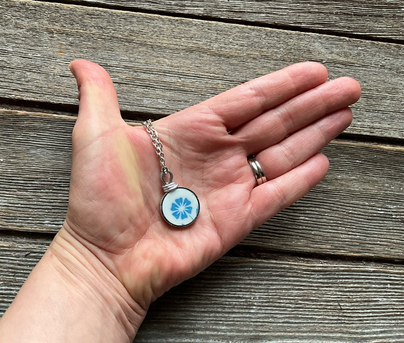 Puede incluir: Un colgante circular con una base de cer&aacute;mica blanca y un patr&oacute;n floral azul, engastado en un marco de metal plateado. El colgante est&aacute; unido a una cadena de eslabones de metal plateado, mostrando un dise&ntilde;o sencillo y delicado.