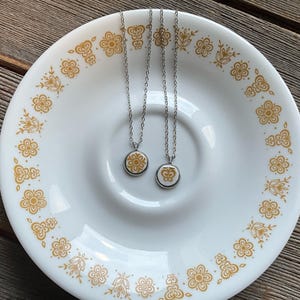 May include: Two silver necklaces with round pendants featuring a gold butterfly design on a white background. The necklaces are displayed on a white plate with a gold floral pattern.