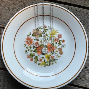 Puede incluir: Cuatro collares de plata con colgantes redondos con flores prensadas. Los colgantes son blancos con un delicado diseño floral en tonos rosa y naranja. Los collares se muestran en un plato blanco con un patrón floral.