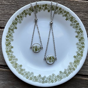 May include: Two silver chain bracelets with adjustable sliding knots. Each bracelet features a round white ceramic charm with a green floral pattern.