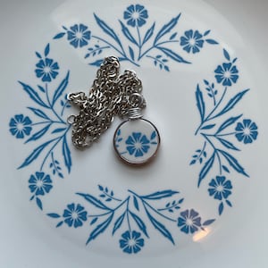 Puede incluir: Un collar con colgante circular que presenta un disco de cer&aacute;mica blanca con un patr&oacute;n floral azul, envuelto en un alambre de tono plateado. El colgante est&aacute; unido a una cadena de eslabones de metal plateado, con un dise&ntilde;o floral azul y blanco.