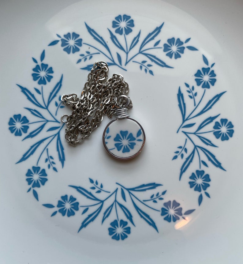 Puede incluir: Un collar con colgante circular que presenta un disco de cer&aacute;mica blanca con un patr&oacute;n floral azul, envuelto en un alambre de tono plateado. El colgante est&aacute; unido a una cadena de eslabones de metal plateado, con un dise&ntilde;o floral azul y blanco.