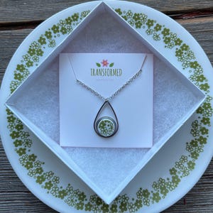 May include: A silver necklace with a teardrop pendant featuring a green floral design. The necklace is in a white box with the text "TRANSFORMED" on the front.