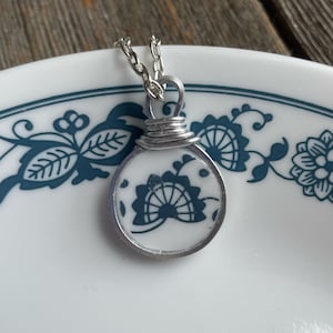 Puede incluir: Un collar con colgante plateado con un colgante redondo blanco con un diseño floral azul. El colgante está encerrado en un marco de metal plateado y cuelga de una fina cadena. El collar se muestra sobre un plato blanco con un borde floral azul.
