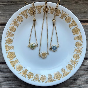 May include: Three gold-toned chain necklaces with circular pendants featuring floral designs in green, blue, and cream colors. The necklaces are displayed on a white plate with a gold floral pattern.